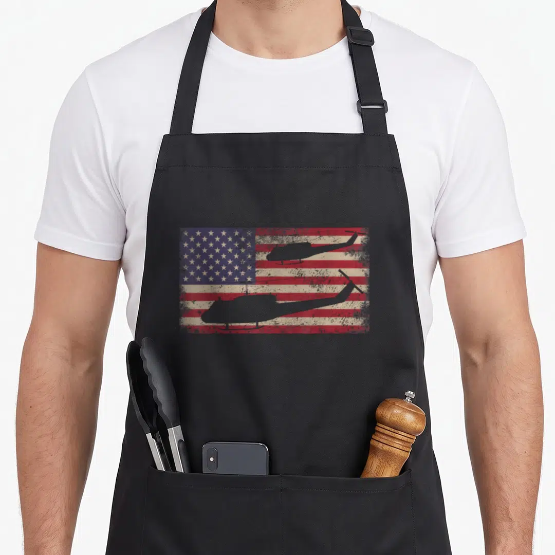 Uh1 american flag apron - Two grey helicopter silhouettes flying over a distressed red white and blue American flag — shown on a black cotton canvas apron against a white background