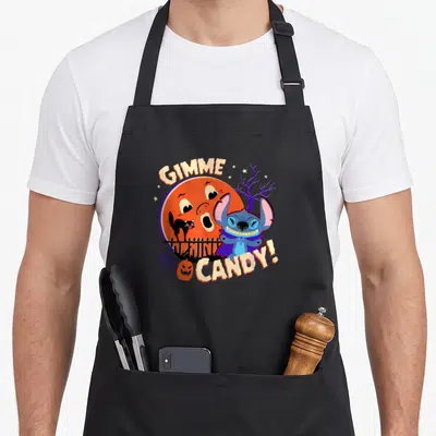 Stitch spooky apron - GIMME CANDY! text around blue character in purple cape, orange moon, cat, pumpkin, fence — shown on a black cotton canvas apron against a white background