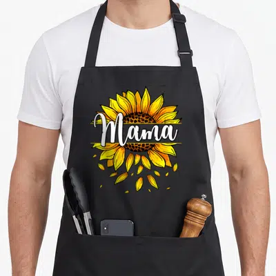 Mama sunflower mother's day aprons - White Mama script over yellow sunflower with brown center and falling petals — shown on a black cotton canvas apron against a white background