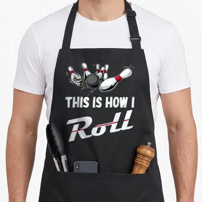 Cute bowling roll apron - Cartoon black ball hits pins with red bands above white text reading THIS IS HOW I Roll — shown on a black cotton canvas apron against a white background