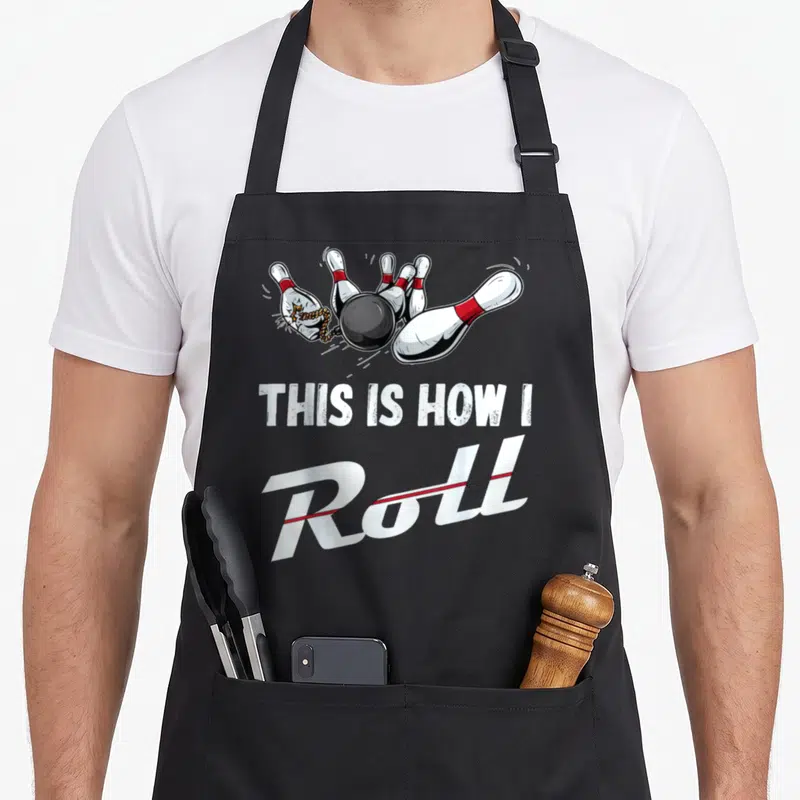 Cute bowling roll apron - Cartoon black ball hits pins with red bands above white text reading THIS IS HOW I Roll — shown on a black cotton canvas apron against a white background