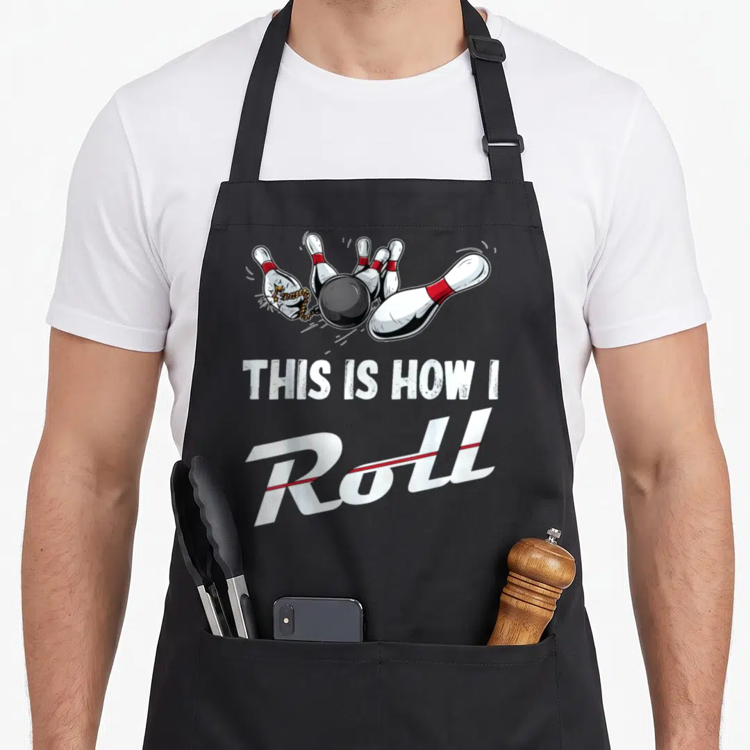 Cute bowling roll apron - Cartoon black ball hits pins with red bands above white text reading THIS IS HOW I Roll — shown on a black cotton canvas apron against a white background