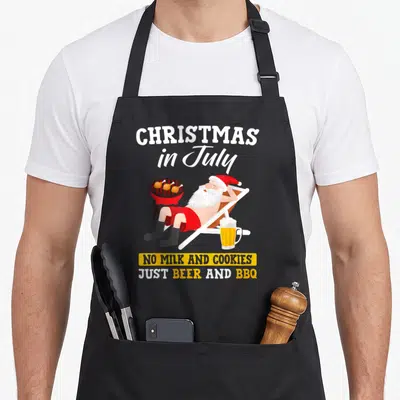 July women's christmas aprons - Santa in shorts by a grill with beer and Christmas in July text — shown on a black cotton canvas apron against a white background