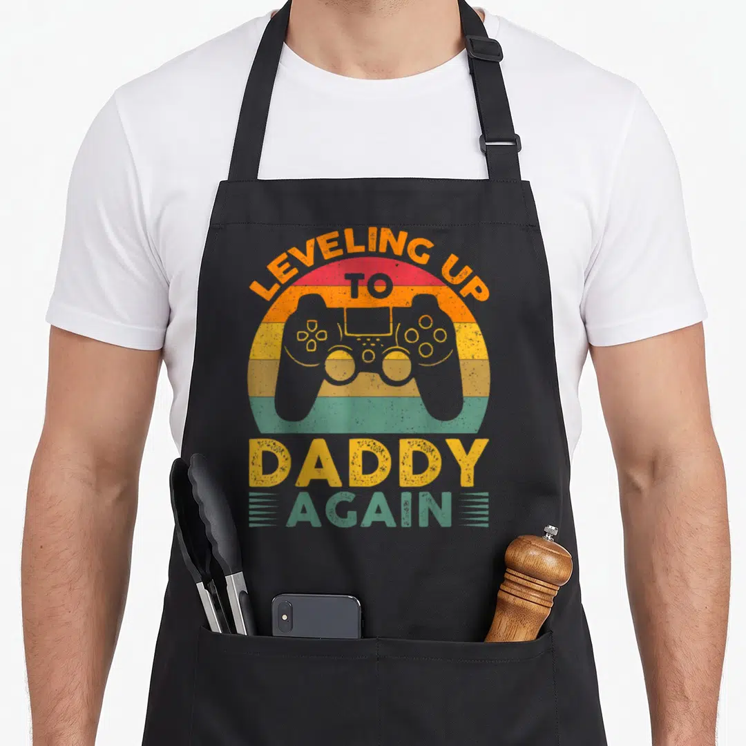 Leveling promoted daddy apron - Grey game controller over striped sunset circle with leveling up to daddy again text — shown on a black cotton canvas apron against a white background