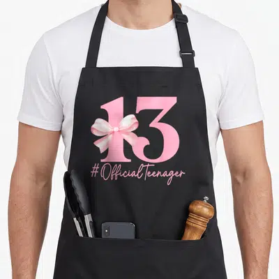 Birthday girly apron - Pink number 13 with satin bow and #OfficialTeenager text on black background — shown on a black cotton canvas apron against a white background