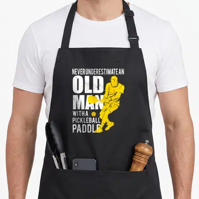 Underestimate paddle pickleball apron - Stacked white text and yellow pickleball player silhouette with ball — shown on a black cotton canvas apron against a white background