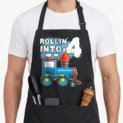 Supply birthday apron - Blue cartoon train with white smoke forming the number 4 and text Rollin Into 4 — shown on a black cotton canvas apron against a white background