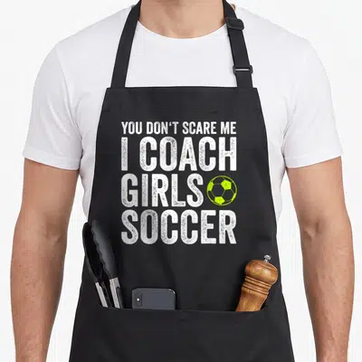 Soccer panic attack apron - Stacked white text You Don't Scare Me I Coach Girls Soccer beside a lime green soccer ball — shown on a black cotton canvas apron against a white background