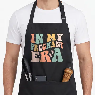 Era pregnant apron - Retro bubbly text In My Pregnant Era with baby footprints and stars in peach teal yellow — shown on a black cotton canvas apron against a white background