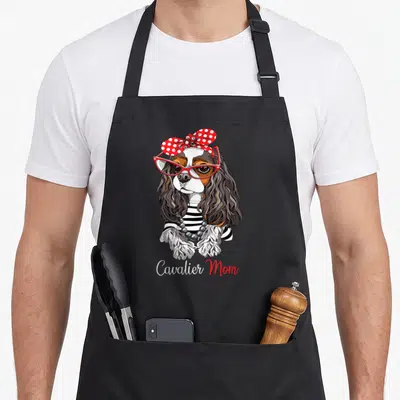 Tricolor king apron - Cartoon tricolor spaniel in red glasses and polka dot bow with Cavalier Mom text — shown on a black cotton canvas apron against a white background