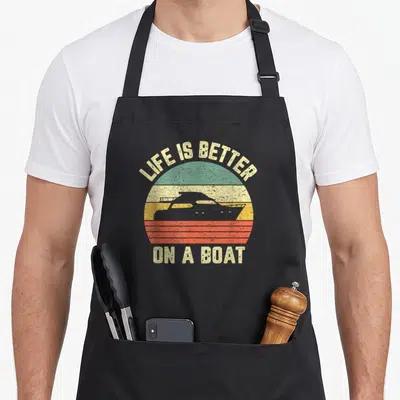 Boating picky apron - Retro striped sun with black boat silhouette and text LIFE IS BETTER ON A BOAT — shown on a black cotton canvas apron against a white background