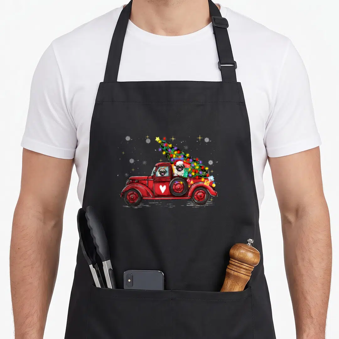 Pug red christmas apron - Red truck carrying two pugs and a decorated Christmas tree with snowflakes — shown on a black cotton canvas apron against a white background