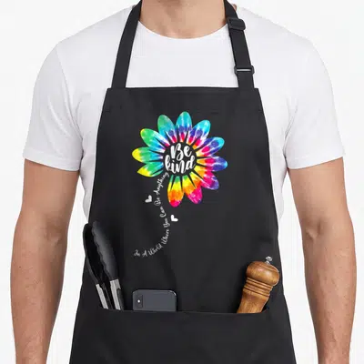 Unity tie-dye apron - Rainbow tie-dye flower with Be Kind text and two hearts on black bib — shown on a black cotton canvas apron against a white background
