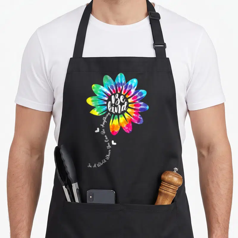 Unity tie-dye apron - Rainbow tie-dye flower with Be Kind text and two hearts on black bib — shown on a black cotton canvas apron against a white background