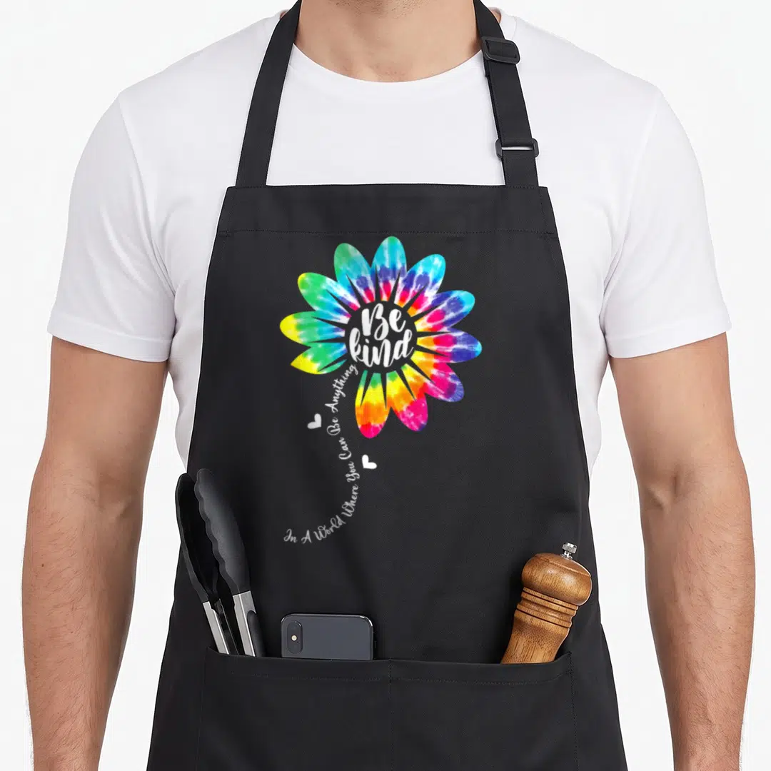 Unity tie-dye apron - Rainbow tie-dye flower with Be Kind text and two hearts on black bib — shown on a black cotton canvas apron against a white background