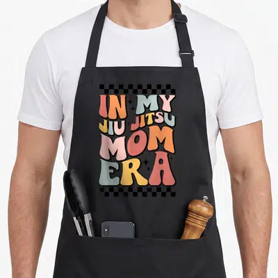 Jiu mom era apron - Retro stacked text In My Jiu Jitsu Mom Era with checkerboard borders in orange blue pink yellow — shown on a black cotton canvas apron against a white background