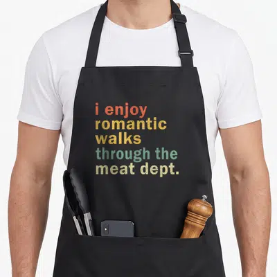 Romantic meat aprons - Stacked colorful text reading i enjoy romantic walks through the meat dept — shown on a black cotton canvas apron against a white background
