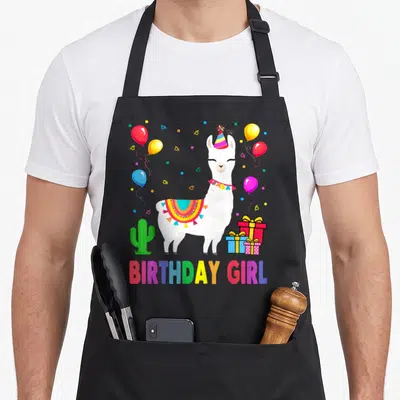 Cactus alpaca apron - White llama wearing party hat and saddle, surrounded by balloons, cactus, gifts, and BIRTHDAY — shown on a black cotton canvas apron against a white background