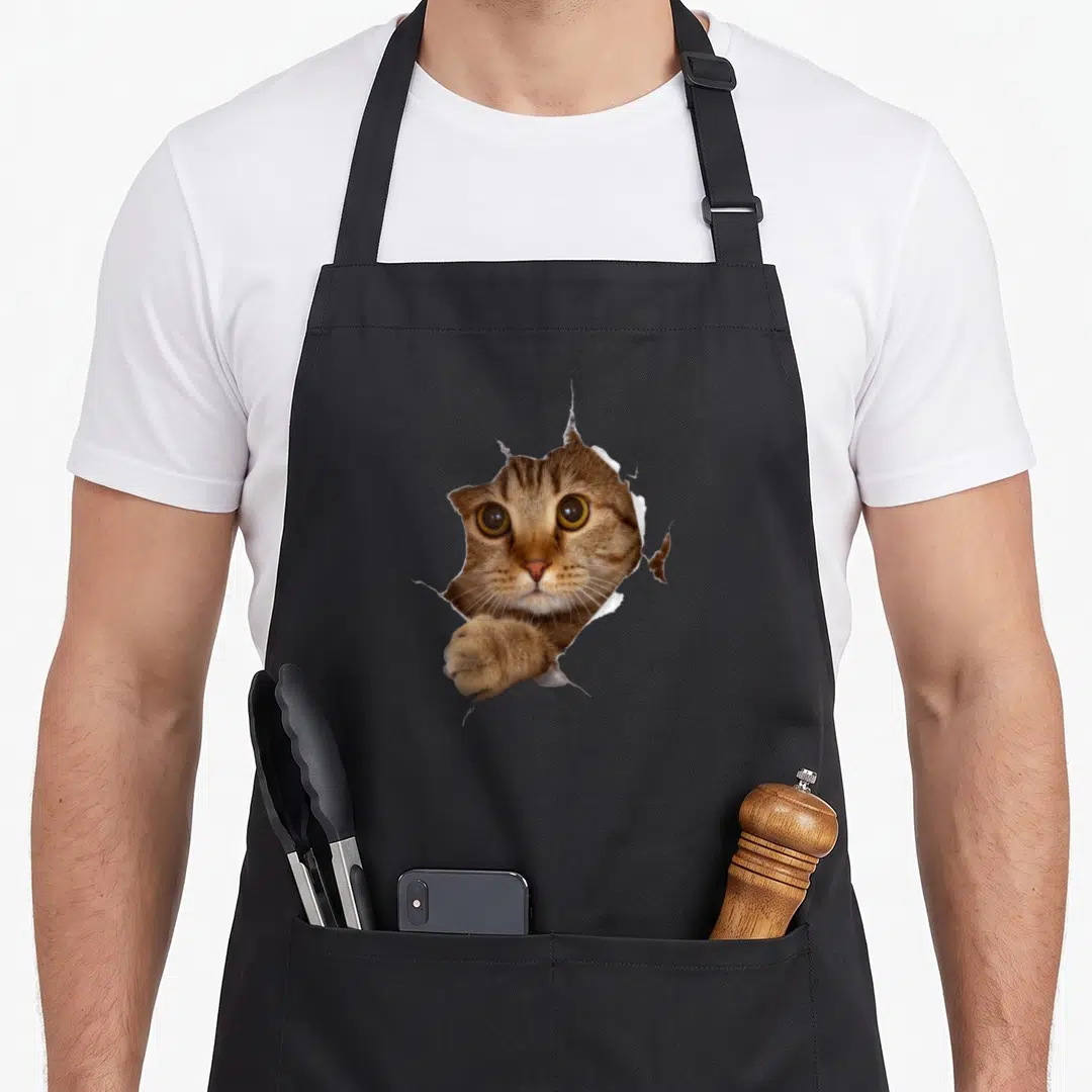 Sweet cat lady apron - Realistic brown and orange cat head and paw breaking through torn white paper — shown on a black cotton canvas apron against a white background
