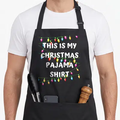 Holiday christmas lights apron - White text THIS IS MY CHRISTMAS PAJAMA SHIRT with colorful light strands — shown on a black cotton canvas apron against a white background