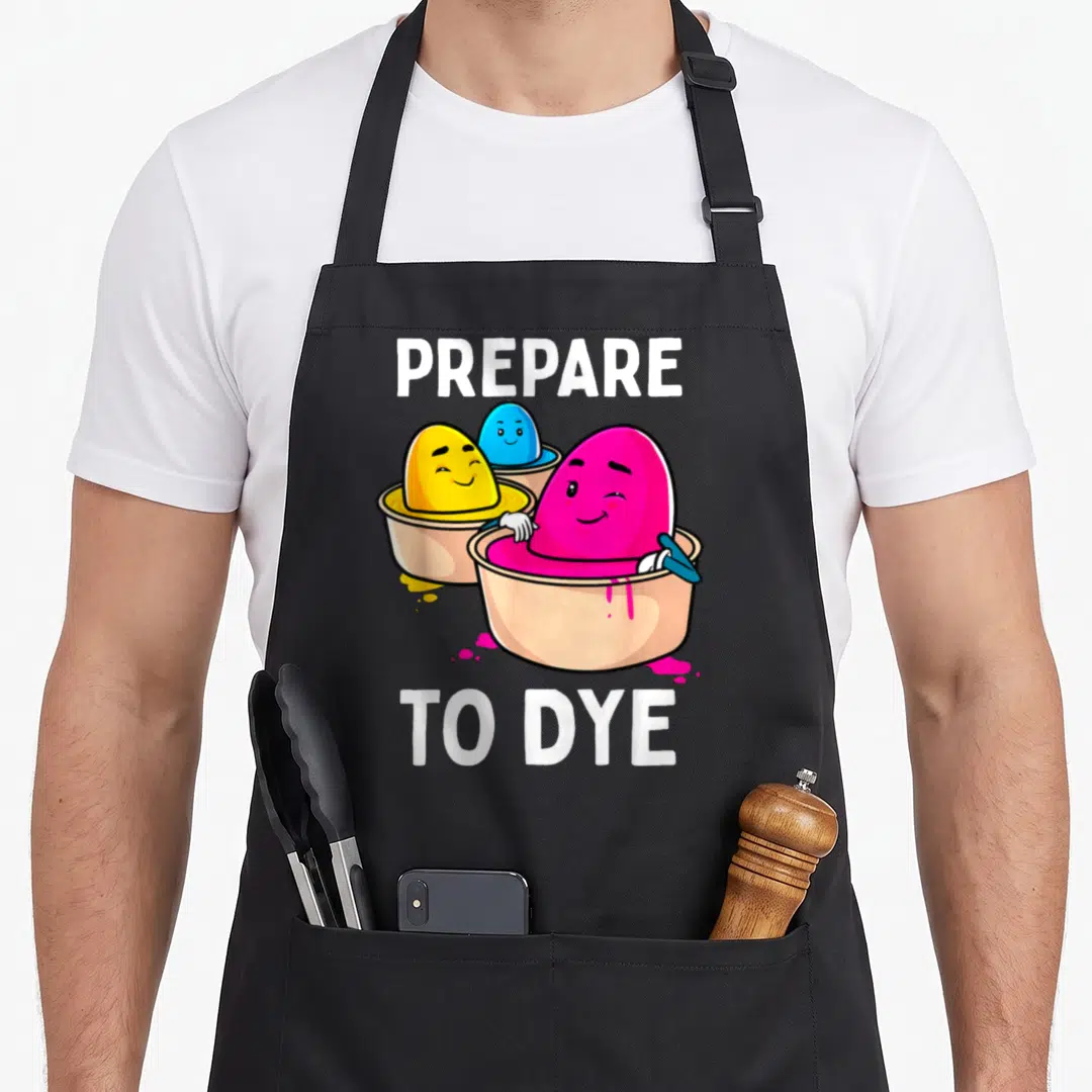 Prepare easter egg apron - PREPARE TO DYE text above three cartoon eggs in pink, yellow, and blue dye bowls — shown on a black cotton canvas apron against a white background
