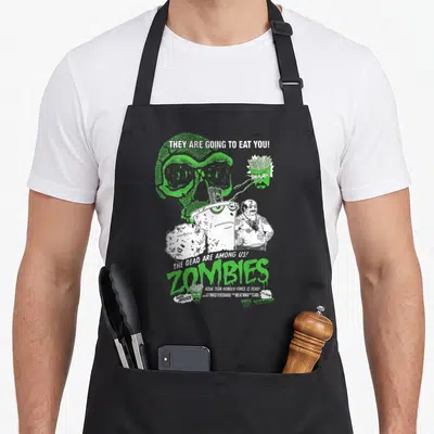 Zomby aqua apron - Green and white cartoon zombies with stacked text and skull graphic — shown on a black cotton canvas apron against a white background