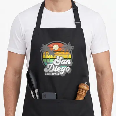 San retro beach vacation summer quotes apron - Retro sunset with palm trees birds and San Diego California text — shown on a black cotton canvas apron against a white background
