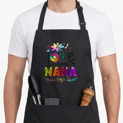 Daisy nana aprons - Rainbow watercolor text love being called NANA with flower and hearts — shown on a black cotton canvas apron against a white background