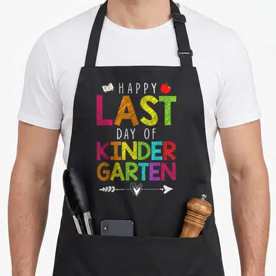 Student kindergarten teacher apron - Stacked HAPPY LAST DAY OF KINDERGARTEN text with book, apple, and heart arrow — shown on a black cotton canvas apron against a white background