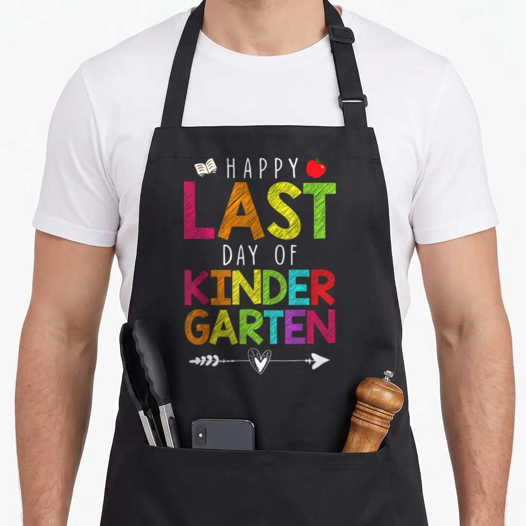 Student kindergarten teacher apron - Stacked HAPPY LAST DAY OF KINDERGARTEN text with book, apple, and heart arrow — shown on a black cotton canvas apron against a white background