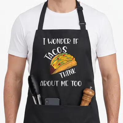 Wonder taco apron - Cartoon taco with yellow, green, red, brown fillings and white text I Wonder If Tacos Think About — shown on a black cotton canvas apron against a white background