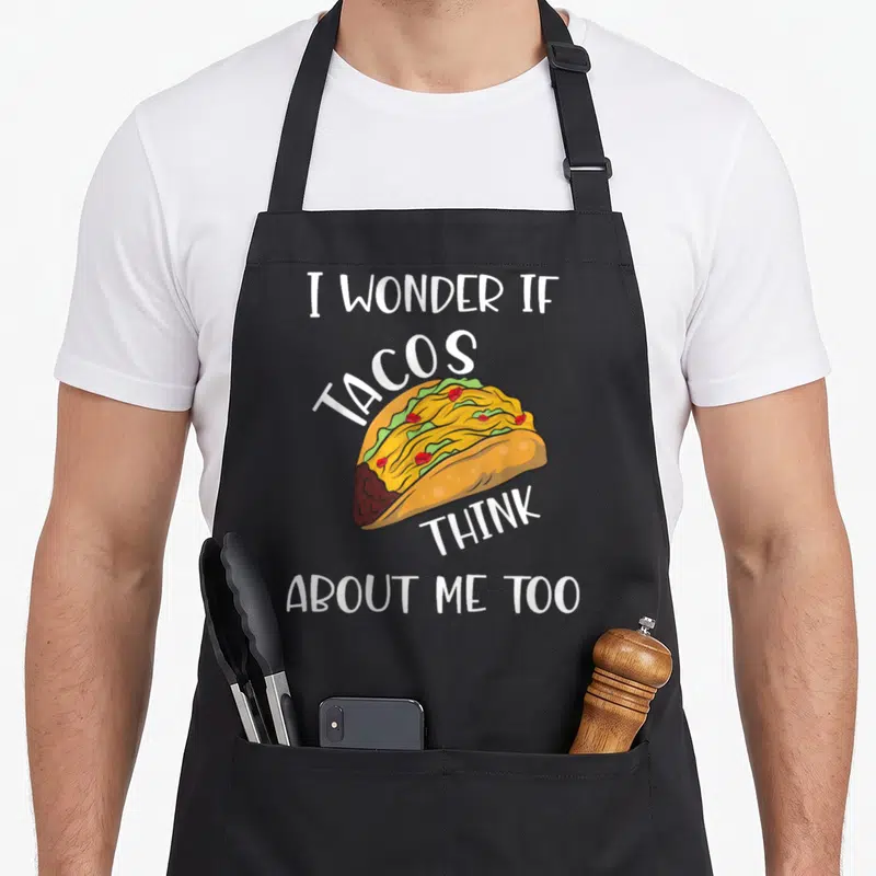 Wonder taco apron - Cartoon taco with yellow, green, red, brown fillings and white text I Wonder If Tacos Think About — shown on a black cotton canvas apron against a white background