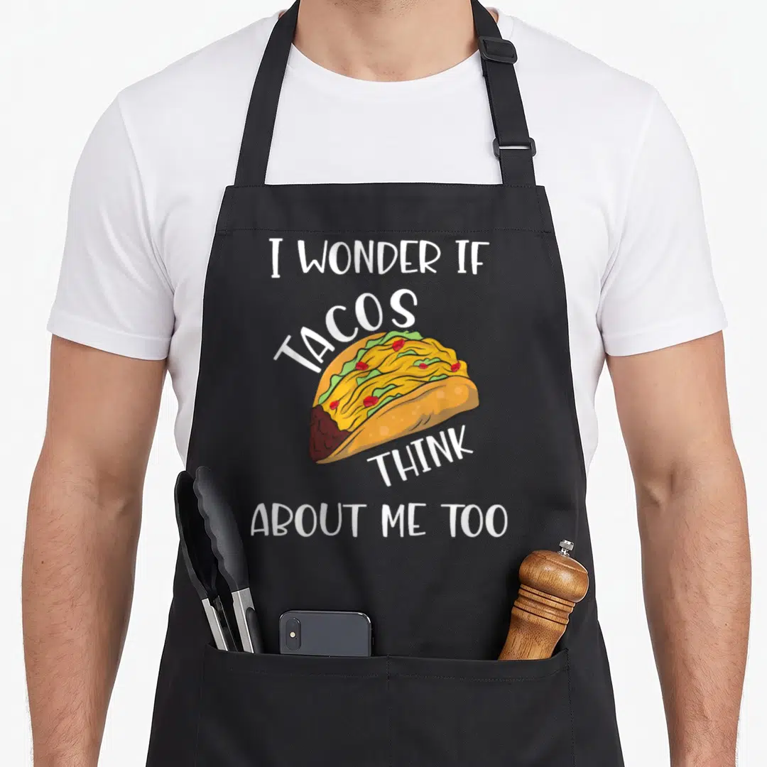 Wonder taco apron - Cartoon taco with yellow, green, red, brown fillings and white text I Wonder If Tacos Think About — shown on a black cotton canvas apron against a white background