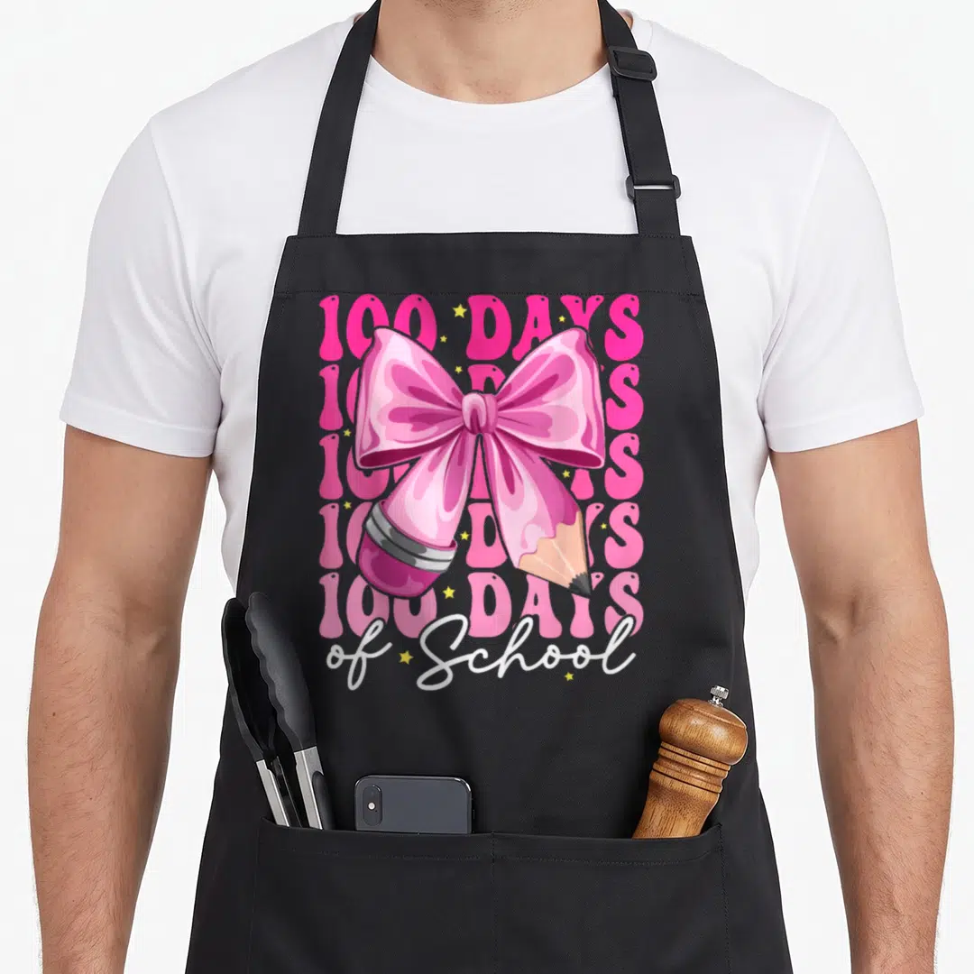 100th bow apron - Pink bow tied to pencil with 100 DAYS text and yellow stars — shown on a black cotton canvas apron against a white background