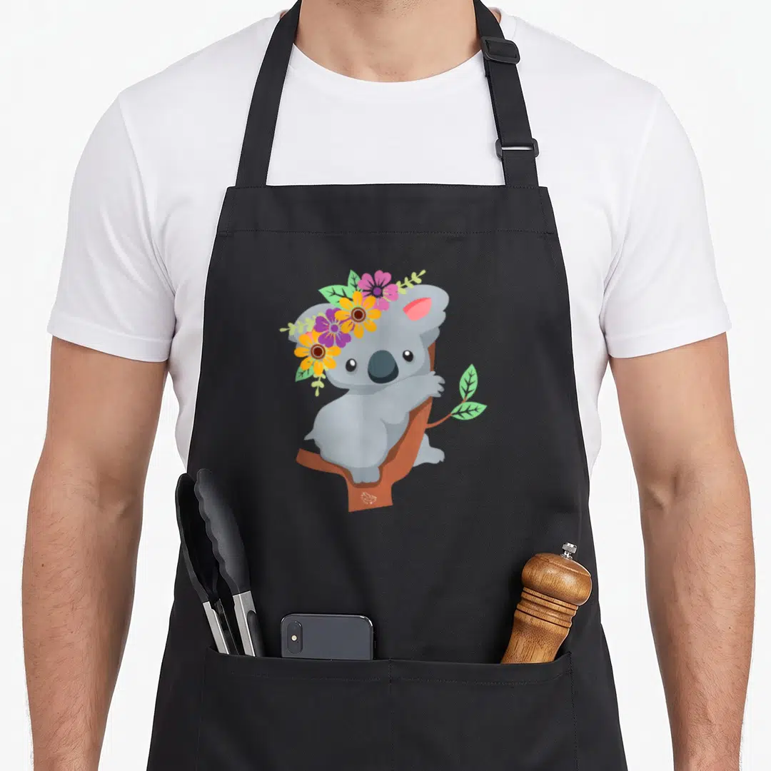 Koala australian apron - Grey cartoon koala in brown tree wearing pink yellow purple flower crown — shown on a black cotton canvas apron against a white background