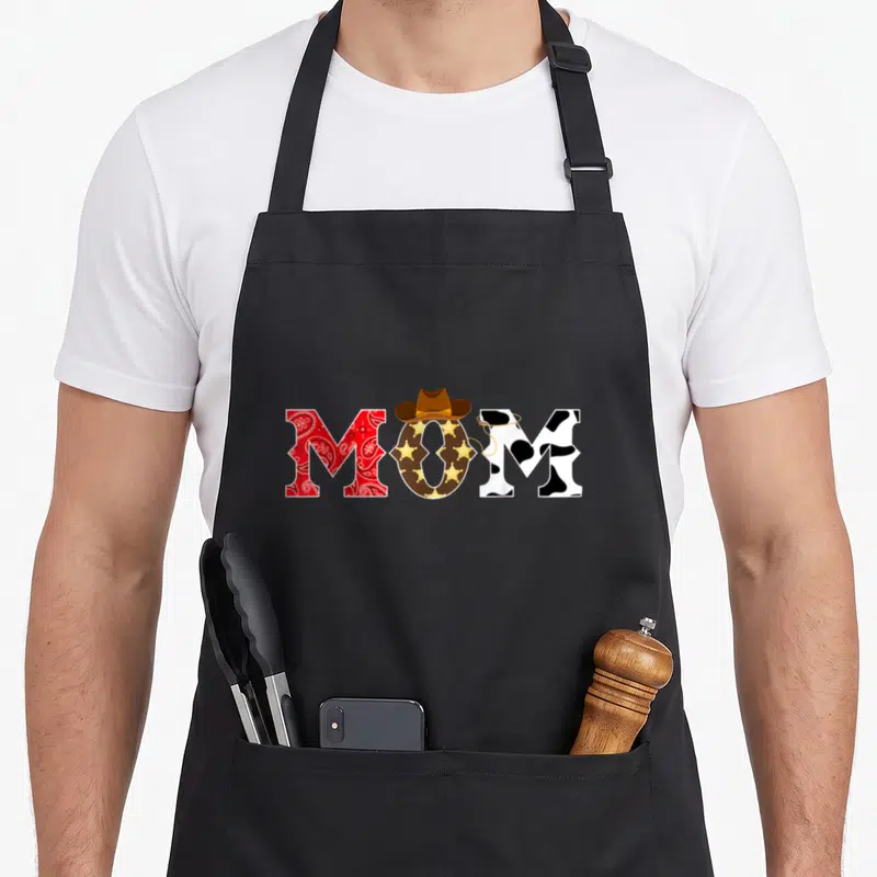 Birthday western aprons - MOM text with cowboy hat, lasso, red paisley, cow print, and sheriff stars — shown on a black cotton canvas apron against a white background