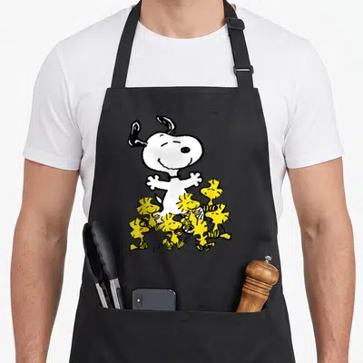 Chick crew apron - White cartoon Snoopy surrounded by cluster of small yellow Woodstock birds on black background — shown on a black cotton canvas apron against a white background