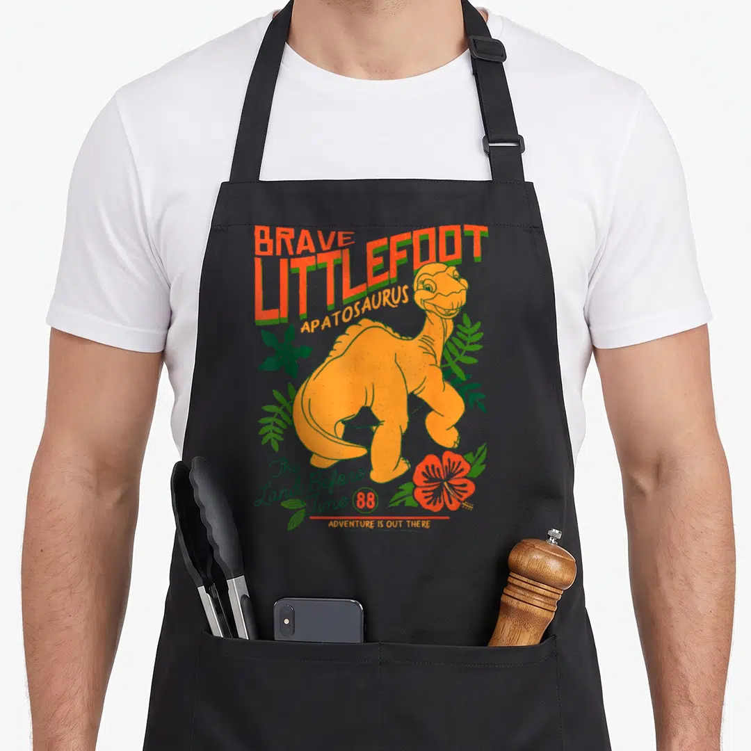 Before novelty apron - Orange Littlefoot dinosaur with green foliage and text The Land Before Time 88 — shown on a black cotton canvas apron against a white background