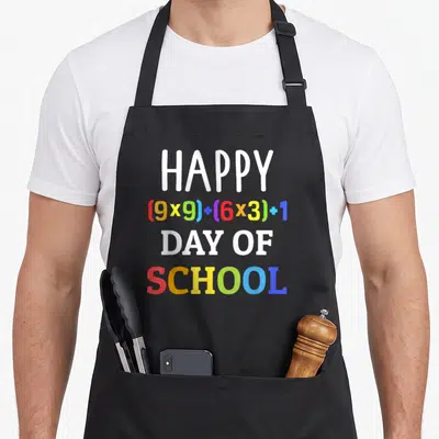 100th formula apron - Stacked text reading HAPPY 9x9 plus 6x3 plus 1 DAY OF SCHOOL in rainbow colors — shown on a black cotton canvas apron against a white background