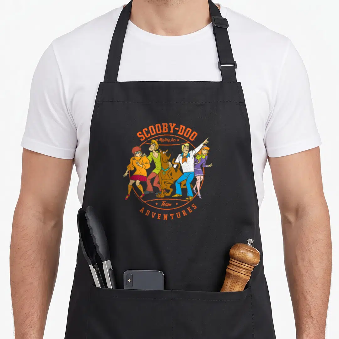 Adventure decade apron - Scooby-Doo gang and Mystery Inc text in orange circular border with cartoon characters — shown on a black cotton canvas apron against a white background