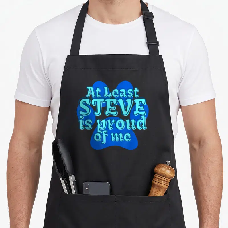 Blue cartoon apron - Blue paw print background with stacked light blue text reading At Least Steve Is Proud Of Me — shown on a black cotton canvas apron against a white background