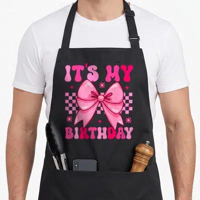 Groovy bow apron - Pink bow over checkerboard with IT'S MY BIRTHDAY text and cartoon stars — shown on a black cotton canvas apron against a white background