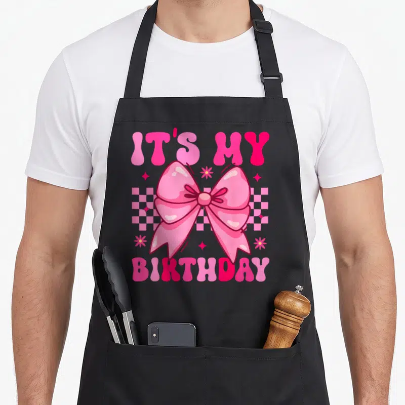 Groovy bow apron - Pink bow over checkerboard with IT'S MY BIRTHDAY text and cartoon stars — shown on a black cotton canvas apron against a white background