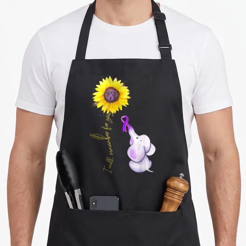 Elephant sunflower apron - Watercolor elephant holding purple ribbon next to sunflower with I will remember for you text — shown on a black cotton canvas apron against a white background