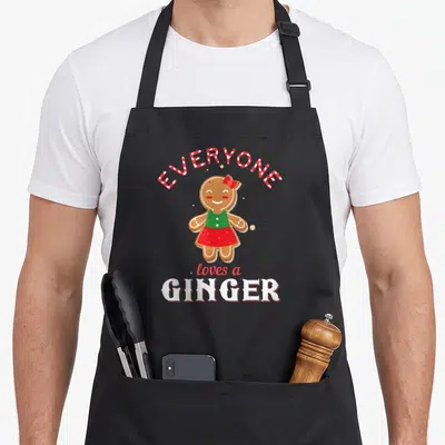 Ginger christmas gingerbread apron - Cartoon gingerbread person in red skirt and green top with arched candy cane text — shown on a black cotton canvas apron against a white background