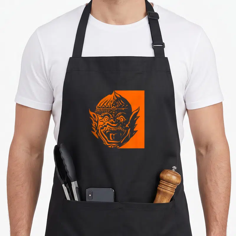 Reamker cambodian apron - Stylized orange demon mask head against a solid orange rectangle on black fabric — shown on a black cotton canvas apron against a white background