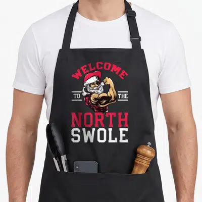 Weightlifting christmas apron - Muscular Santa flexing under Welcome to the North Swole text in red and white — shown on a black cotton canvas apron against a white background