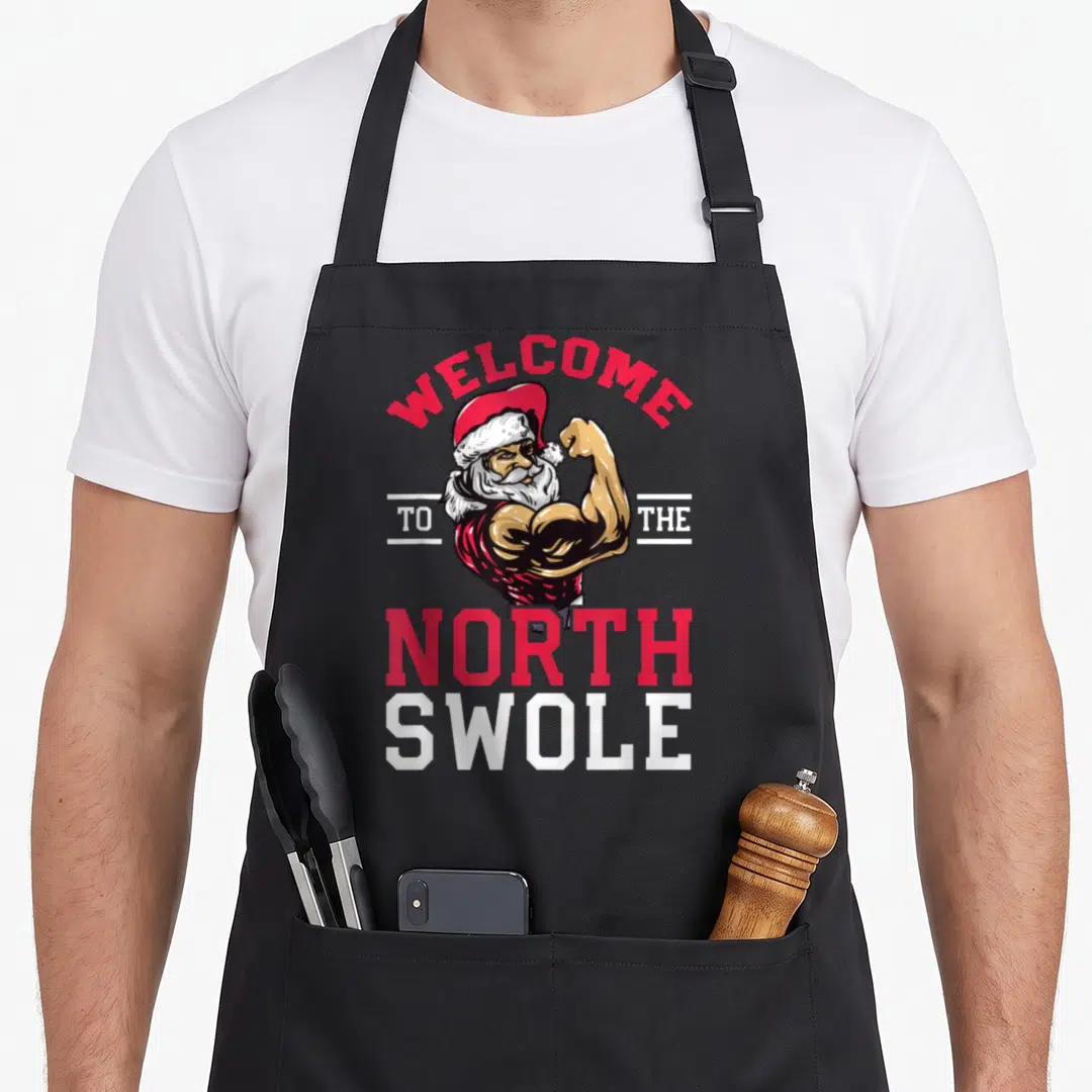 Weightlifting christmas apron - Muscular Santa flexing under Welcome to the North Swole text in red and white — shown on a black cotton canvas apron against a white background