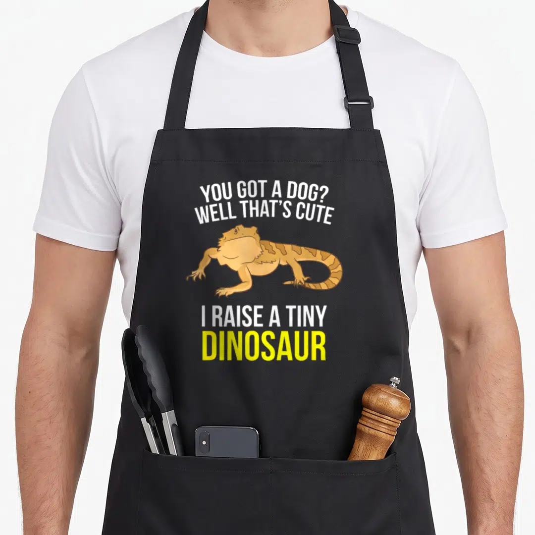 Dragon custom pet apron - Bearded dragon illustration between text You Got A Dog Well That's Cute I Raise A Tiny — shown on a black cotton canvas apron against a white background