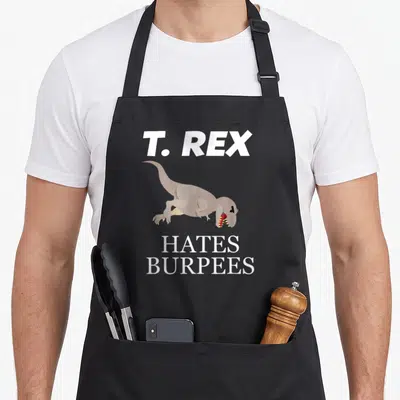 Hate t-rex apron - Cartoon tan T-Rex with white text T. REX HATES BURPEES — shown on a black cotton canvas apron against a white background